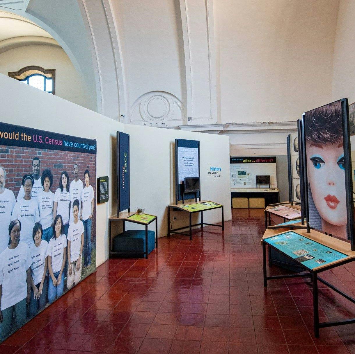 An image inside the "Race: Are We So Different?" exhibit featuring a life-sized group portraits of diverse people on the left, and various tables and museum signage, one depicting a large image of a white children's doll, on the right.