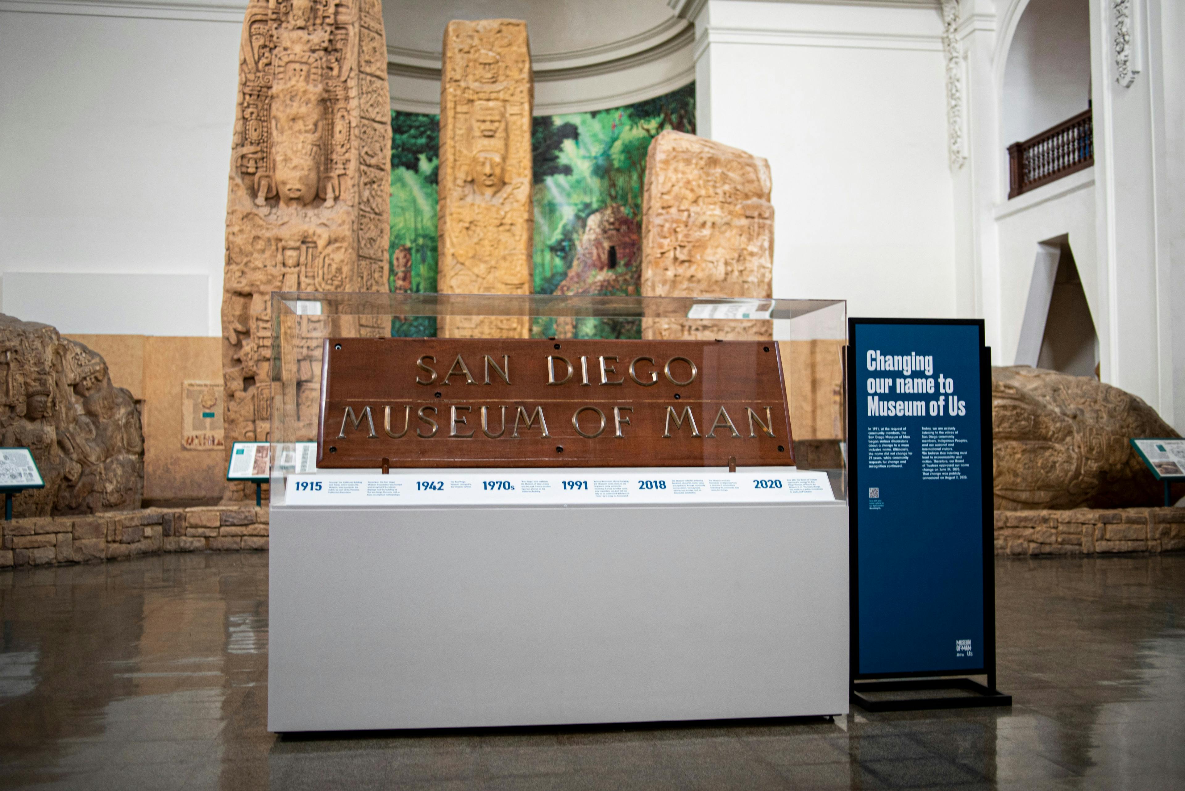 The wooden "San Diego Museum of Man" sign with brass letters sits within a plexiglass case. Underneath it, is a timeline with historic dates for the museum. To the right a blue standing sign reads, "Changing our name to Museum of Us." The whole installation stands before the Museum's "Maya Peoples: Heart of Sky, Heart of Earth" exhibit and three Maya stelae monuments.