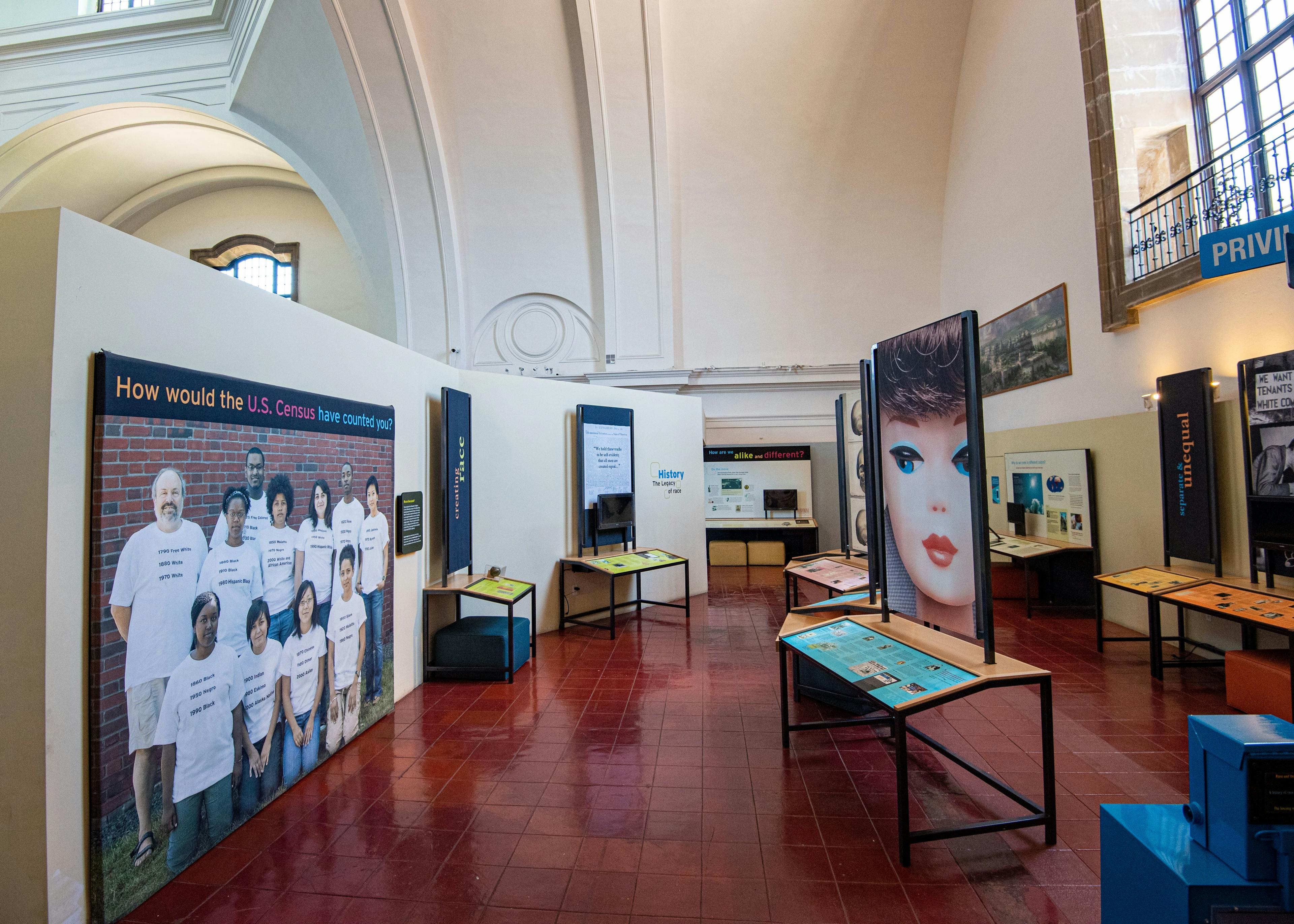 An image inside the "Race: Are We So Different?" exhibit featuring a life-sized group portraits of diverse people on the left, and various tables and museum signage, one depicting a large image of a white children's doll, on the right.