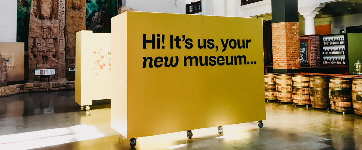 A yellow wall installation that reads "Hi! It's us, your new museum..." in black text.