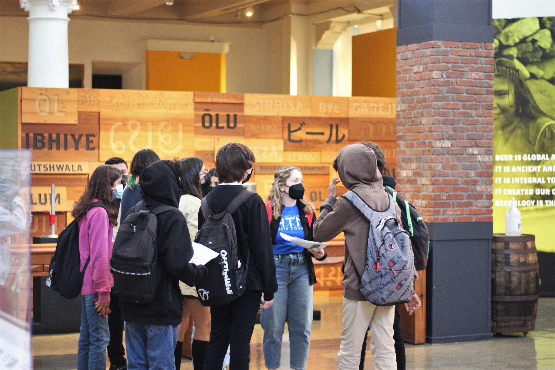 A group of students wearing backpacks in the Museum's rotunda facing towards an instructor in a blue shirt and wearing a black mask.