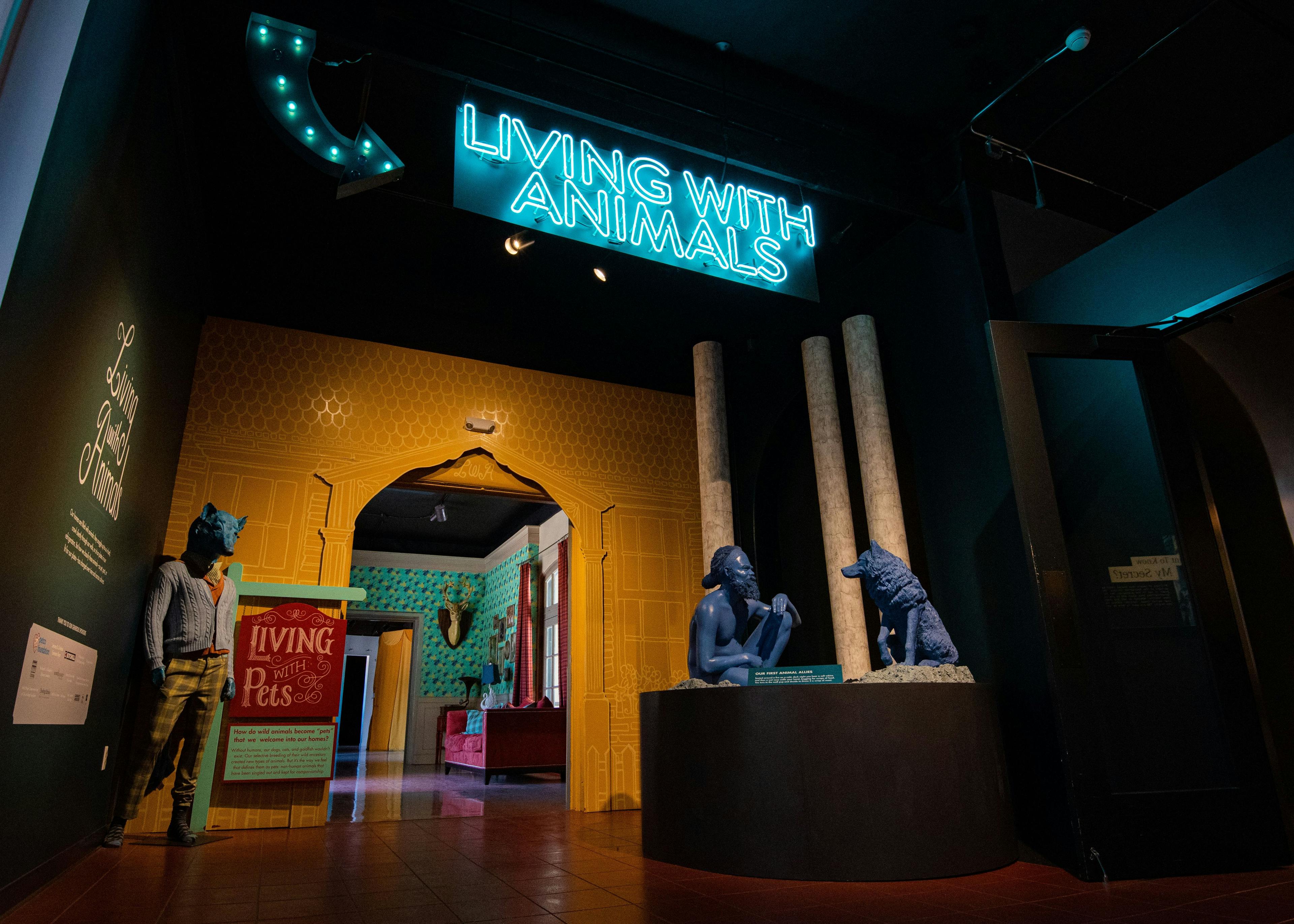 The entrance to the "Living with Animals" exhibit. A neon sign reads the exhibit title. Below it, an ancient homosapien crouches to face a wolf on the left. On the right, a human-like figure sculpture wears a paper macheted fox head. 