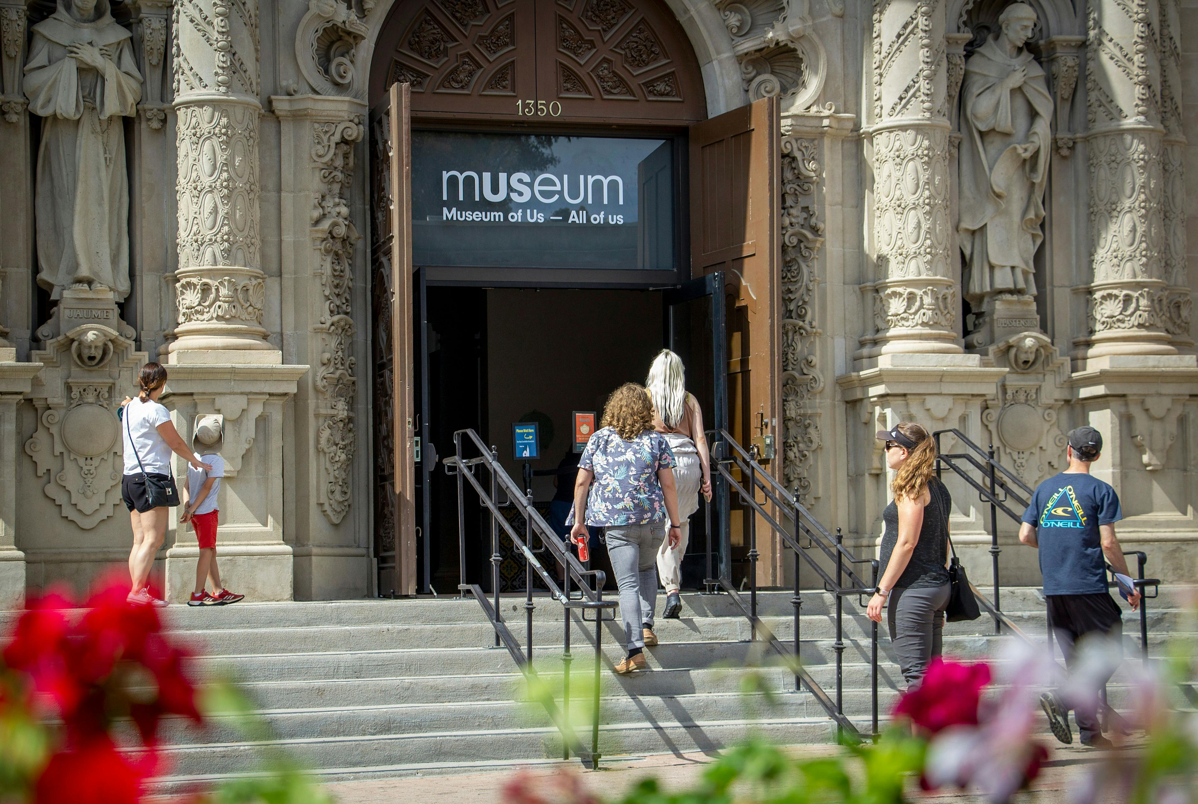 Various people walking up the stairs and into the Museum of Us lobby entrance. In the foreground are potted flowers.