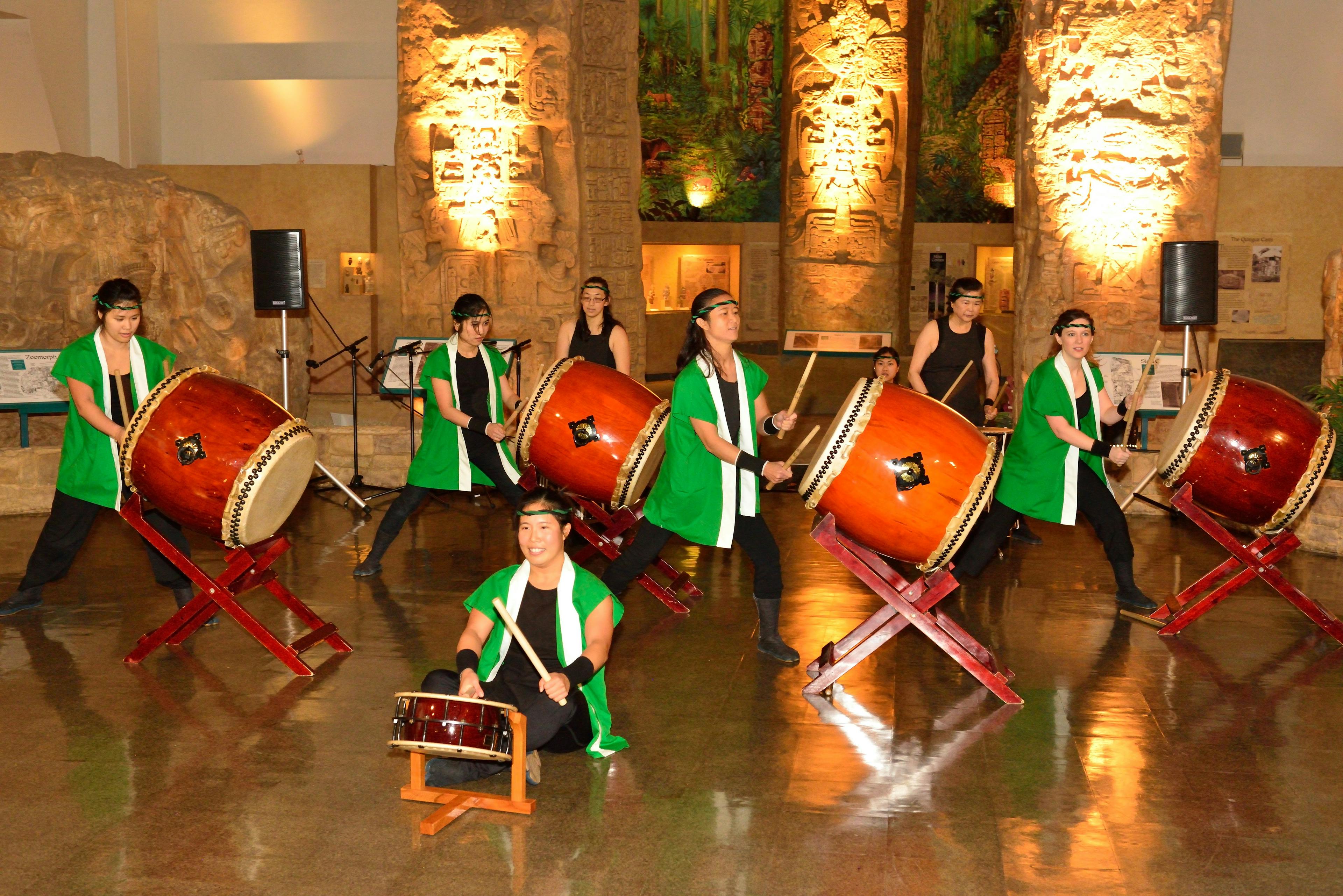 A group of five drummers in green and black garments perform in the Museum rotunda in front of the Maya Peoples exhibit.