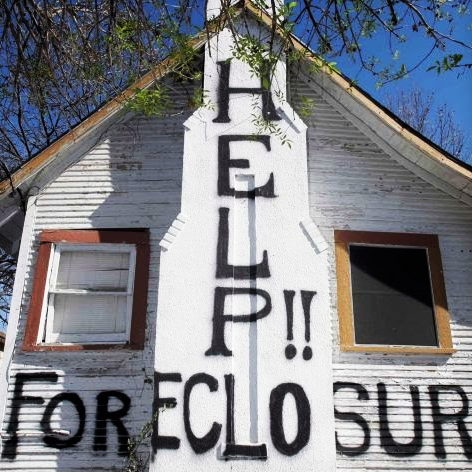 A white house with the windows boarded up with the words "HELP!! FORECLOSURE" painted on the exterior.