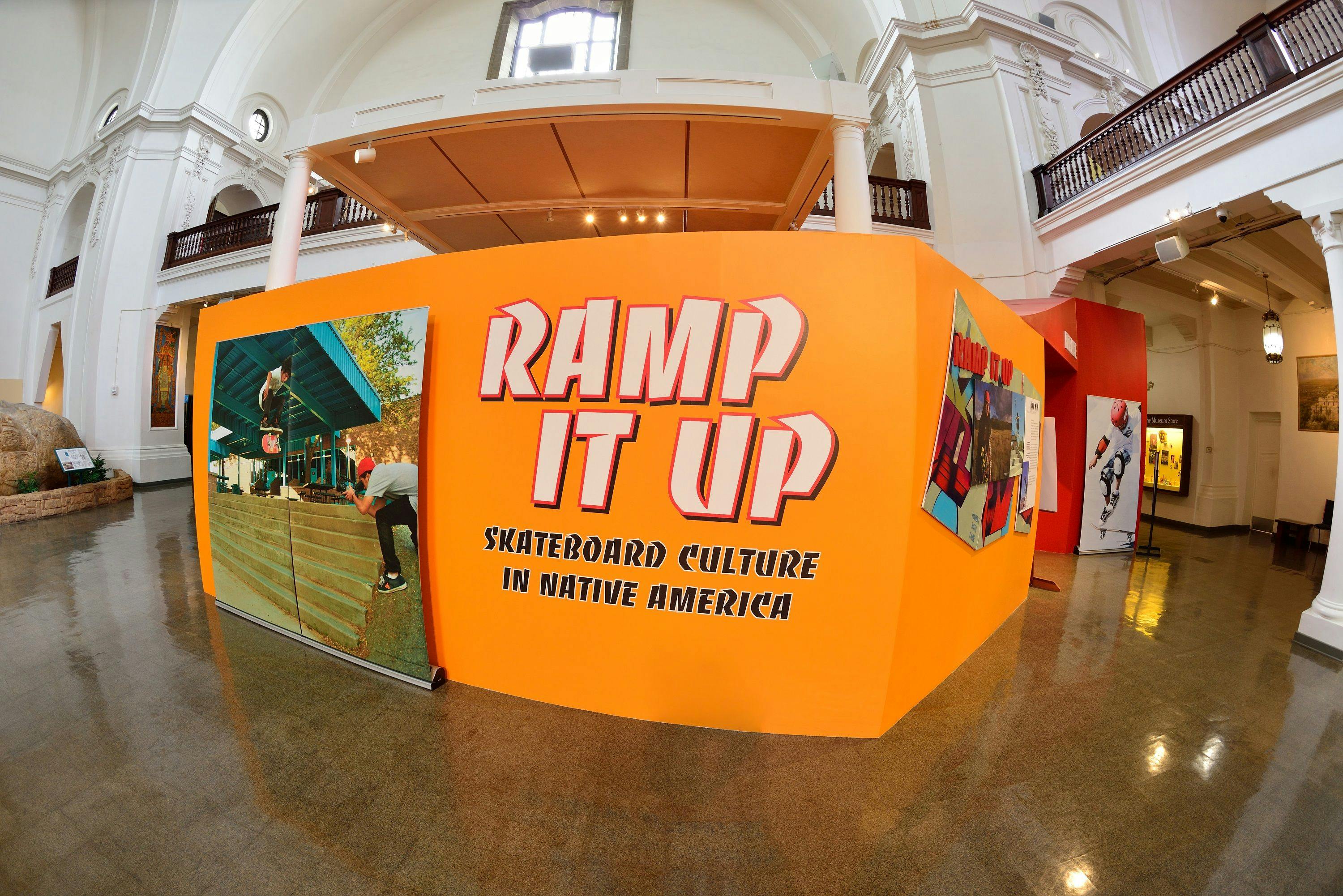 A bright orange exhibit title wall featuring an image of a skater jumping off stairs next to text that reads, "Ramp It Up - Skateboard Culture in Native America"