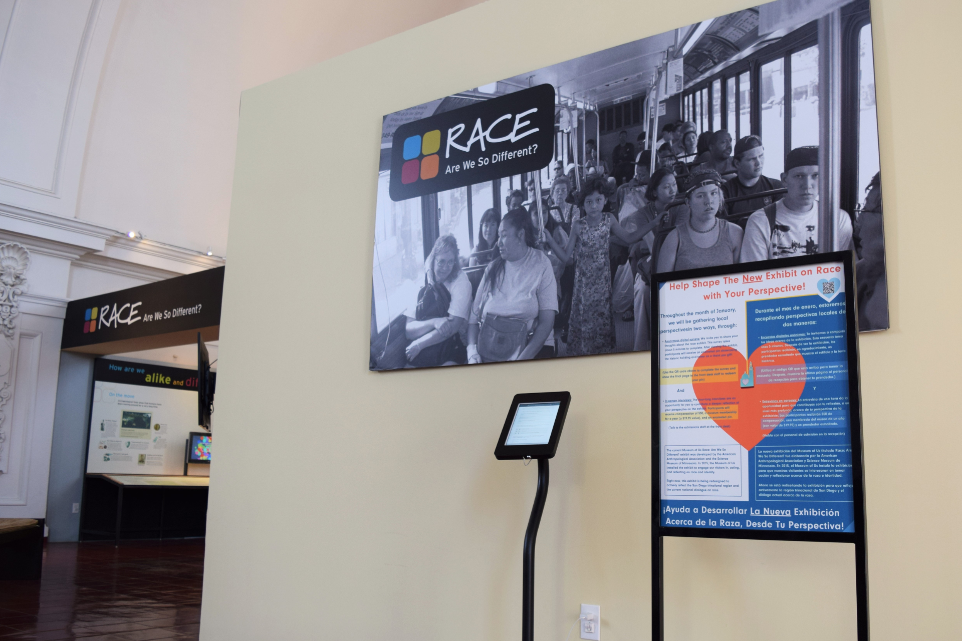 The entry wall of the "Race: Are We So Different?" exhibit, with a large black and white image of several people on a public transit bus centered on the exhibit wall. A large sign next to a small digital kiosk sits right in front of the wall and black and white image. At the top of the large white sign reads, "Help Shape The New Exhibit on Race with Your Perspective!" above two columns of text explaining the exhibit surveys in both English and Spanish.