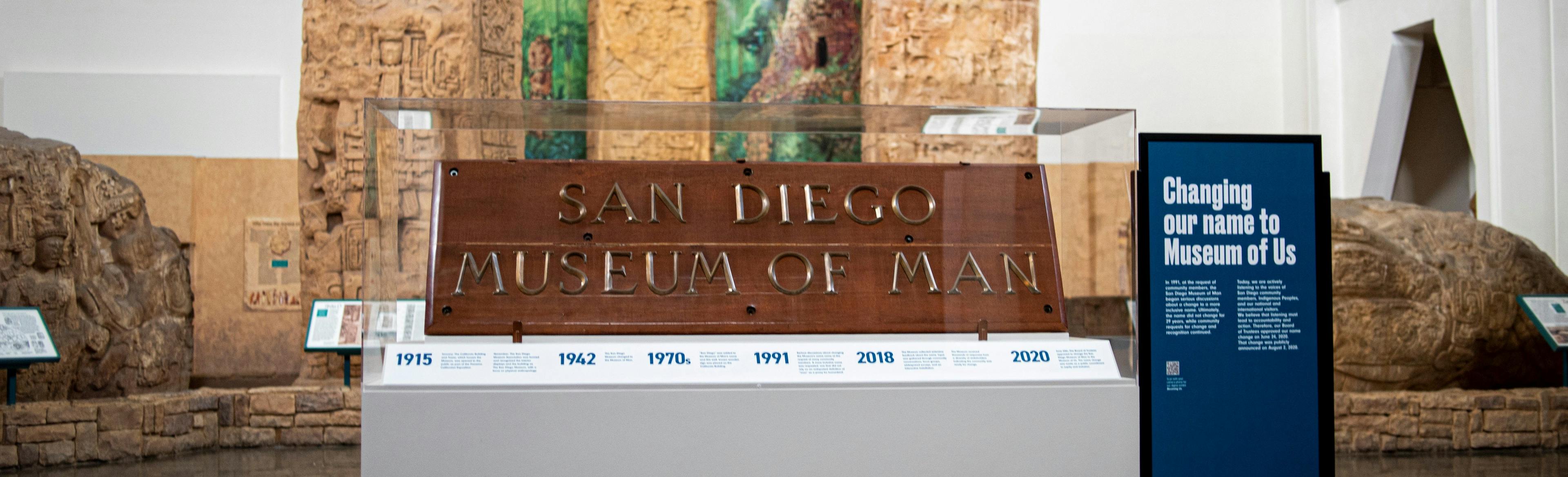 The wooden "San Diego Museum of Man" sign with brass letters sits within a plexiglass case. Underneath it, is a timeline with historic dates for the museum. To the right a blue standing sign reads, "Changing our name to Museum of Us." The whole installation stands before the Museum's "Maya Peoples: Heart of Sky, Heart of Earth" exhibit and three Maya stelae monuments.