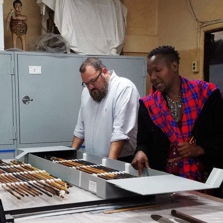 Maasai Cultural Ambassador Ole Sankale examines and discusses a row of spears in a box on a table, as Museum staff listen.
