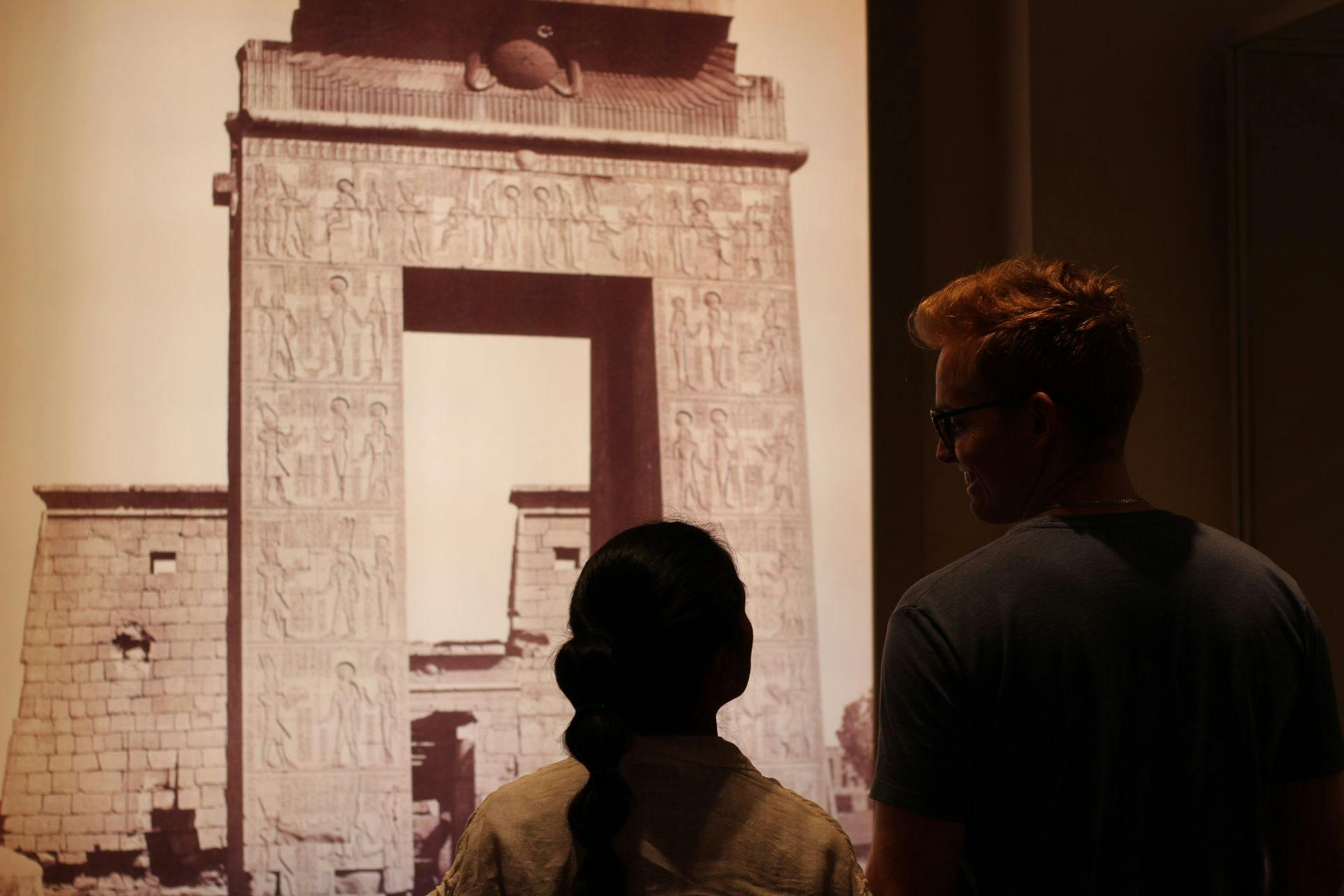 Two people look at each other before an illuminated image of an archway in Ancient Egypt.