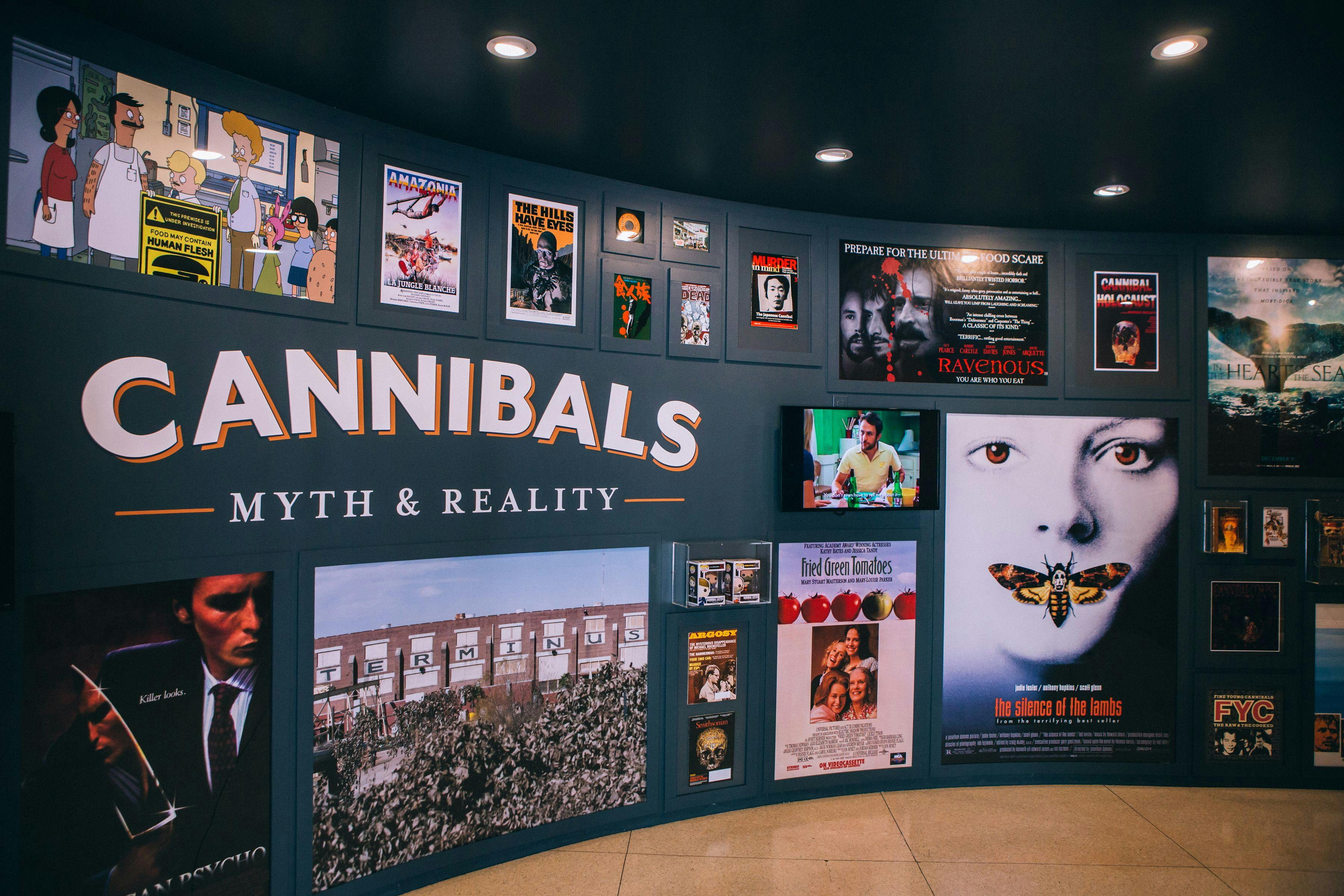 Graphic installation of various movie posters and prints hung on an exhibition wall titled 'Cannibals: Myth & Reality'.