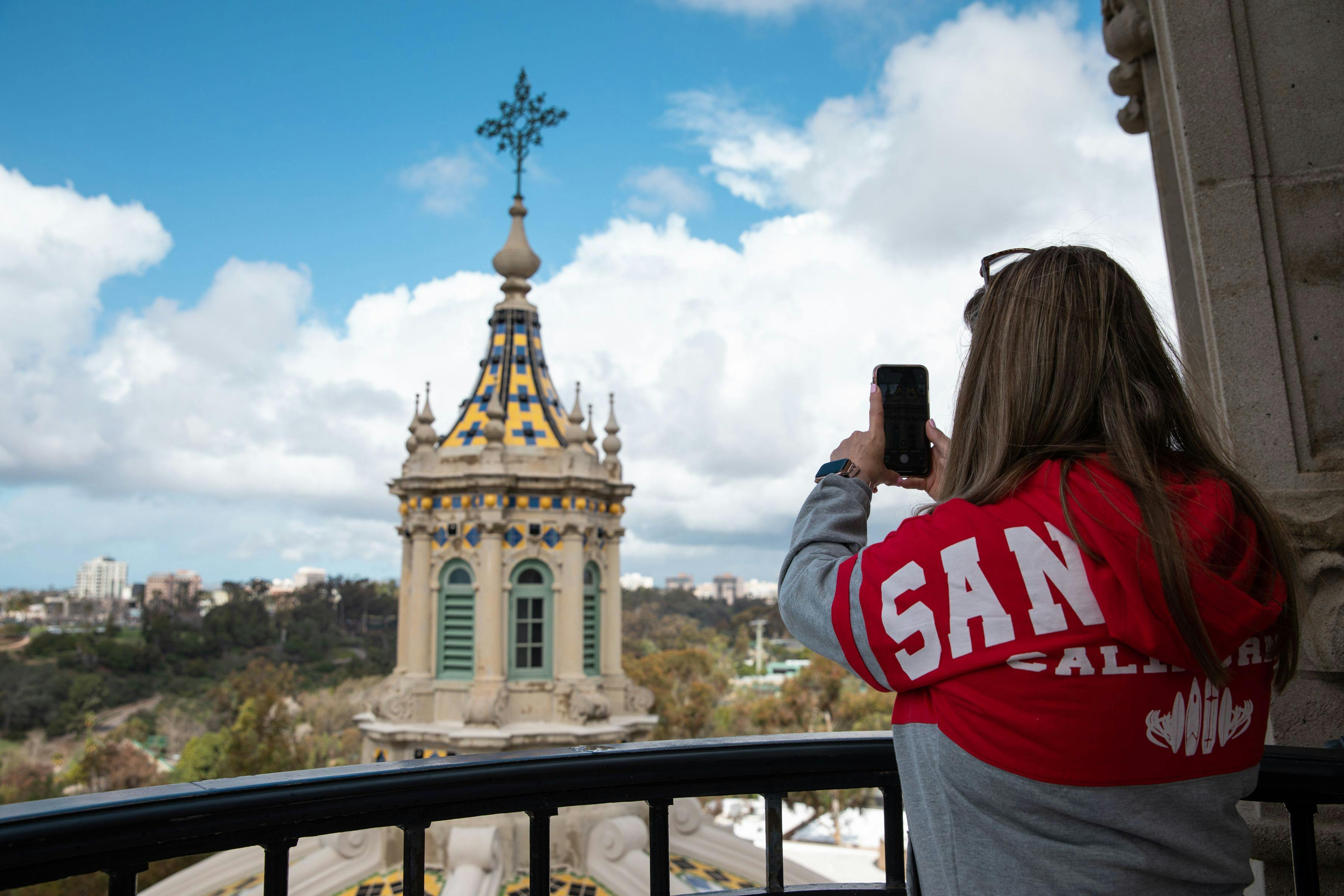 A person in a red and gray long sleeve shirt and long hair takes a picture on their smart phone of the California Building's tiled dome from the California Tower's viewing deck. Foliage and the San Diego skyline is in the background.
